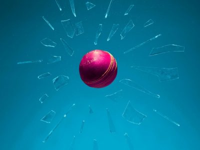 Dynamic shot of a red cricket ball breaking through glass against a teal backdrop.